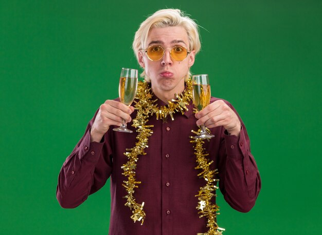 Бестолковый молодой блондин в очках с гирляндой из мишуры на шее держит два бокала шампанского, изолированные на зеленой стене