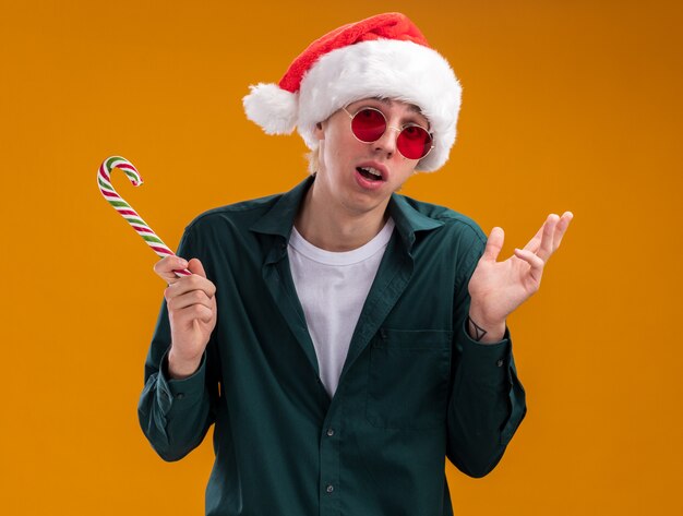 Ignaro giovane uomo biondo che indossa cappello da babbo natale e occhiali che tengono la canna da zucchero natalizia guardando la telecamera che mostra la mano vuota isolata su sfondo arancione