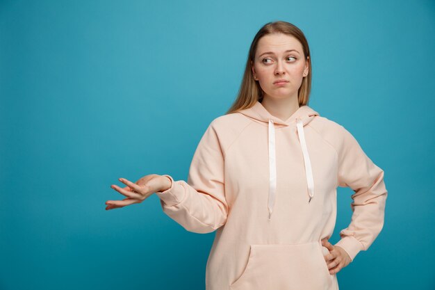 Clueless young blonde woman looking at side keeping hand on waist showing empty hand 