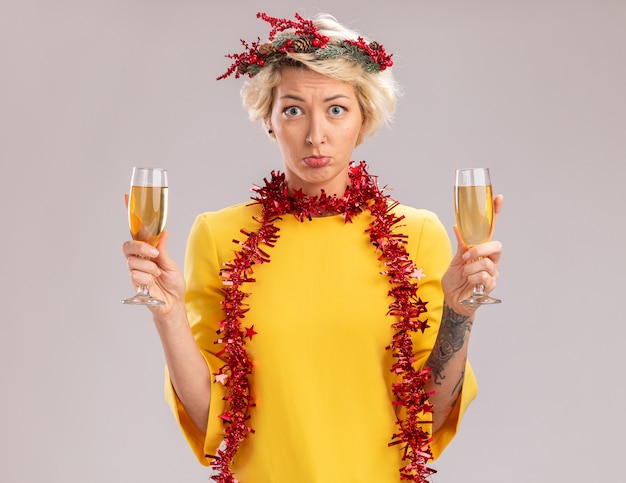 Clueless young blonde woman wearing christmas head wreath and tinsel garland around neck holding two glasses of champagne looking at camera isolated on white background