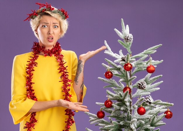 クリスマス ヘッド リースと見掛け倒しの花輪を身に着けている無知な若いブロンドの女性が飾られたクリスマス ツリーの近くに立って、紫の壁に孤立して見える手でそれを指しています。