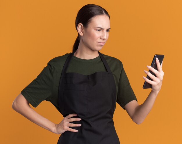 Clueless young brunette barber girl in uniform holds and looks at hair clippers