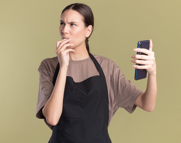 Clueless young brunette barber girl in uniform puts hand on chin and holds phone looking at side on olive green