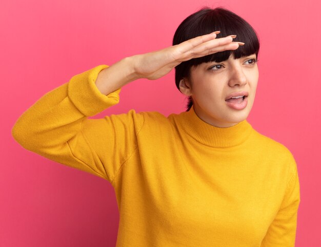 Clueless young brunette caucasian girl keeps palm at forehead looking at side on pink