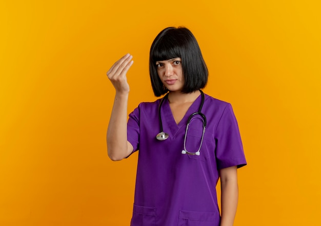 Clueless young brunette female doctor in uniform with stethoscope gestures money hand sign isolated on orange background with copy space