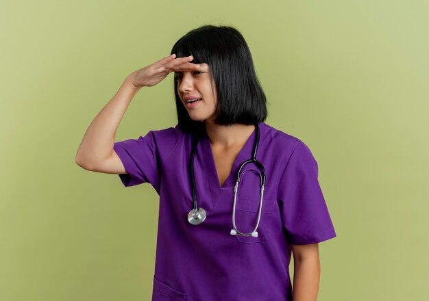 Невежественная молодая брюнетка женщина-врач в униформе со стетоскопом держит ладонь у лба, пытаясь увидеть, глядя в сторону, изолированную на оливково-зеленом фоне с копией пространства