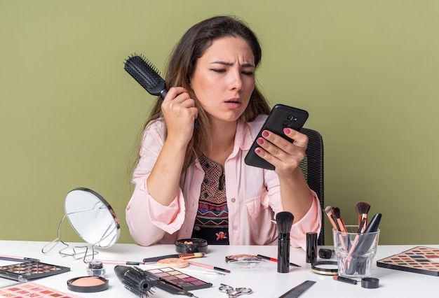 Clueless young brunette girl sitting at table with makeup tools holding comb and looking at phone isolated on olive green wall with copy space