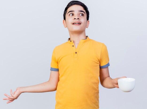 Clueless young caucasian boy looking at side showing empty hand holding cup isolated on white background