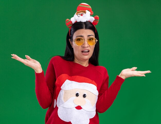 Clueless young caucasian girl wearing santa claus headband and sweater with glasses looking at camera showing empty hands isolated on green background