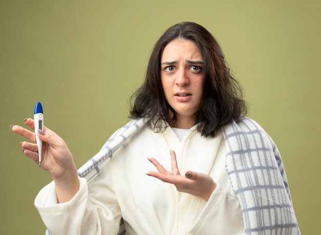 Clueless young caucasian ill girl wearing robe wrapped in plaid showing thermometer pointing at it with hand looking at camera isolated on olive green background