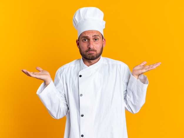 Бесплатное фото Бестолковый молодой кавказский повар в униформе и кепке шеф-повара смотрит в камеру и делает жест, изолированный на оранжевой стене