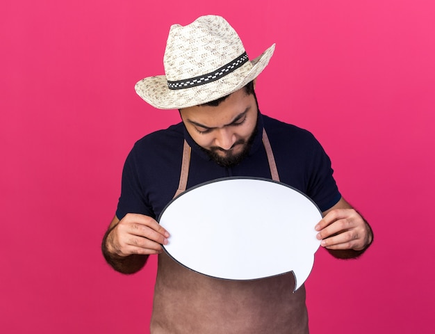 clueless young caucasian male gardener wearing gardening hat holding and looking at speech balloon isolated on pink wall with copy space