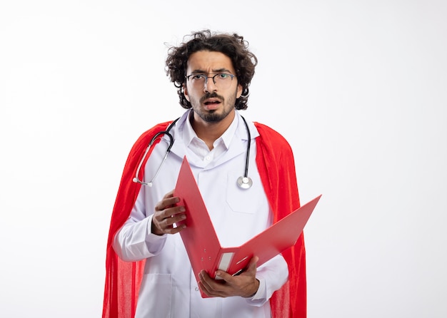 Foto gratuita ignaro giovane supereroe caucasico uomo in occhiali ottici che indossa l'uniforme del medico con mantello rosso e con uno stetoscopio intorno al collo tiene la cartella