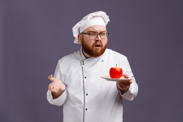 Clueless young chef wearing glasses uniform and cap holding apple in plate looking at side showing empty hand isolated on purple background