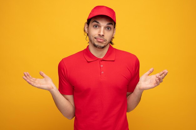 clueless young delivery man wearing uniform and cap looking at camera showing empty hands isolated on yellow background