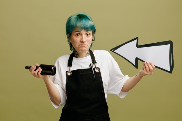 Clueless young female barber wearing uniform holding arrow sign pointing at herself and hair spray looking at camera isolated on olive green background