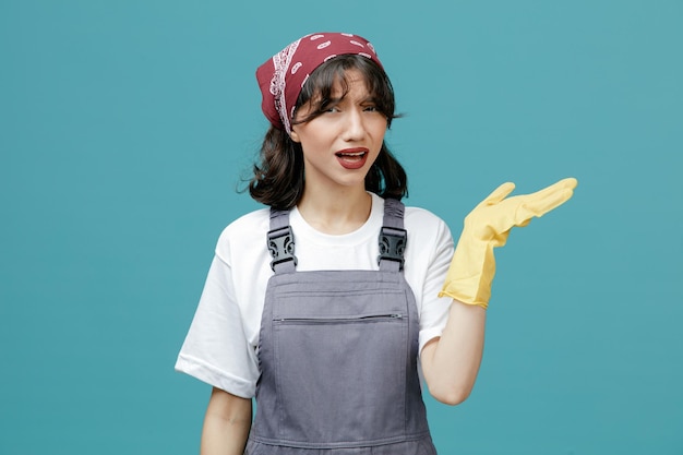 Giovane donna addetta alle pulizie che indossa bandana uniforme e guanti di gomma guardando la fotocamera che mostra la mano vuota isolata su sfondo blu