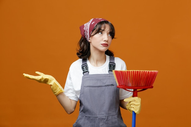 Clueless young female cleaner wearing uniform rubber gloves and bandana holding squeegee mop looking at side showing empty hand isolated on orange background