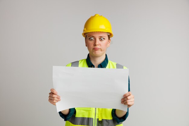 Clueless young female construction worker wearing safety helmet and safety vest holding and looking at paper with pursed lips 