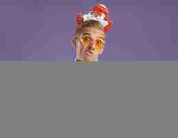 無料写真 サンタクロースのヘッドバンドを身に着けている無知な若いハンサムな男とコピースペースで紫色の壁に分離されたクリスマスボールの飾りを保持している眼鏡とネクタイ