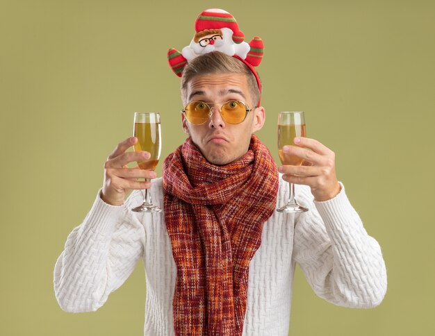 Бестолковый молодой красивый парень в повязке на голову санта-клауса и шарфе с двумя бокалами шампанского, изолированными на оливково-зеленой стене