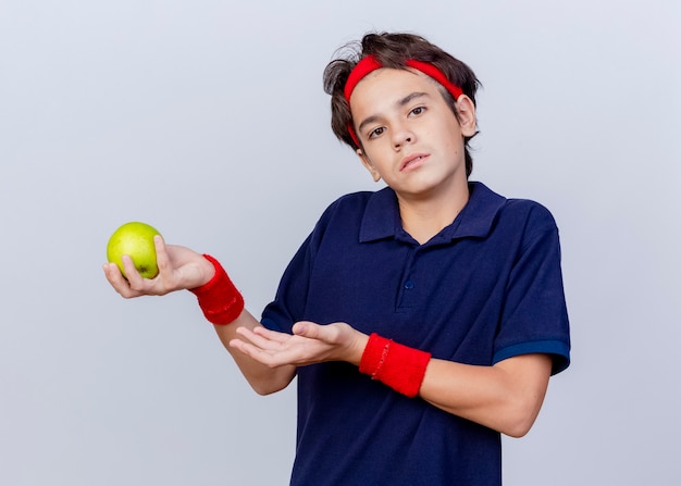 Clueless young handsome sporty boy wearing headband and wristbands with dental braces  holding and pointing with hand at apple isolated on white wall