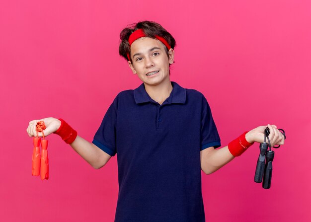 clueless young handsome sporty boy wearing headband and wristbands with dental braces looking at camera holding jump ropes isolated on crimson background