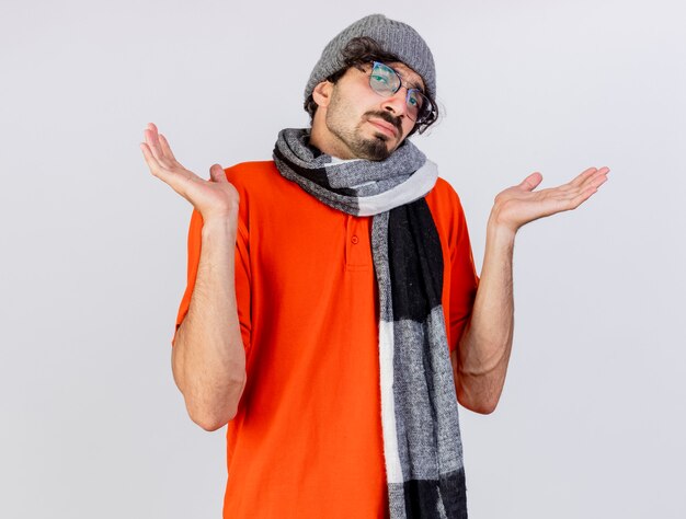 眼鏡の冬の帽子と白い壁に隔離された空の手を示す正面を見てスカーフを身に着けている無知な若い病気の男
