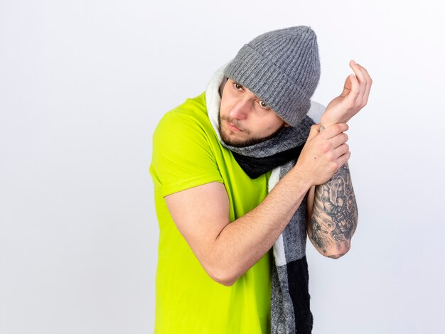 Clueless young ill man wearing winter hat and scarf holds hand trying to hear pulse isolated on white wall