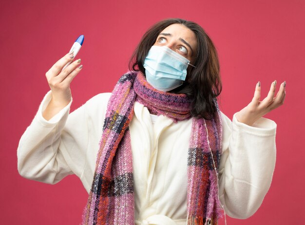 Clueless young ill woman wearing robe and scarf with mask holding thermometer showing empty hand looking up isolated on pink wall