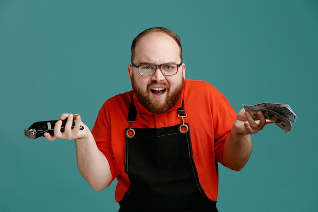 Free photo clueless young male barber wearing glasses red shirt and barber apron looking at camera with money and hair clipper in hands isolated on blue background