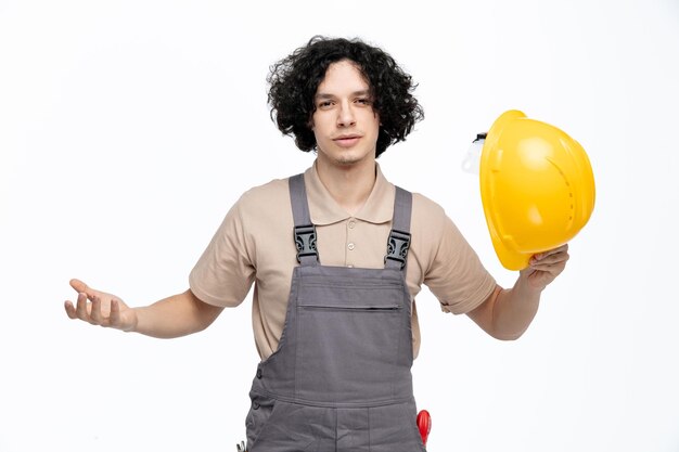 Clueless young male construction worker wearing uniform holding safety helmet looking at camera showing empty hand with construction instruments in his pocket isolated on white background