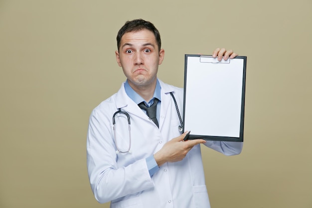 Clueless young male doctor wearing medical robe and stethoscope around neck holding clipboard and pen looking at camera showing clipboard isolated on olive green background