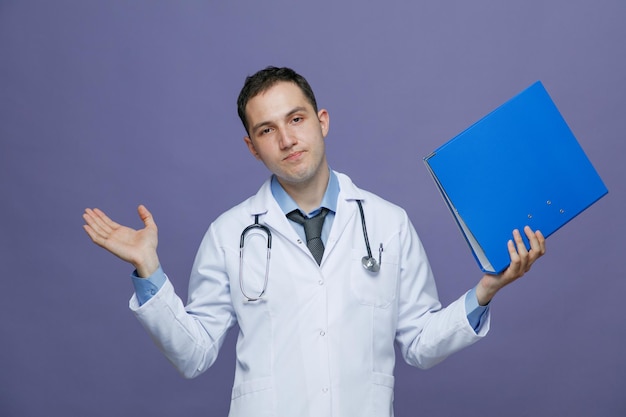 Clueless young male doctor wearing medical robe and stethoscope around neck looking at camera showing folder and empty hand isolated on purple background