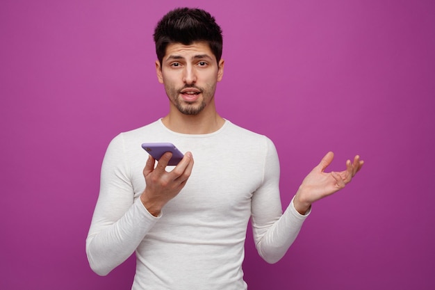 Free photo clueless young man holding mobile phone looking at camera showing empty hand isolated on purple background