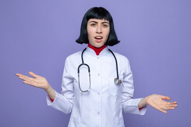 Clueless young pretty caucasian girl in doctor uniform with stethoscope keeping hands open 