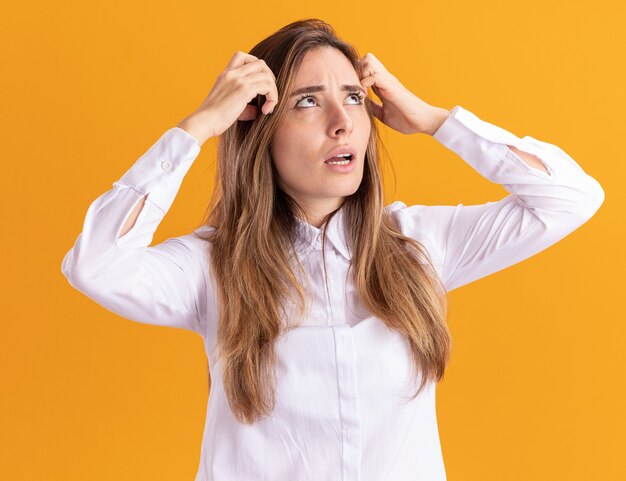 Clueless young pretty caucasian girl puts hands on head and looks up on orange 