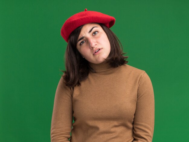 La giovane ragazza abbastanza caucasica senza indizi con il cappello del berretto esamina la macchina fotografica isolata sulla parete verde con lo spazio della copia