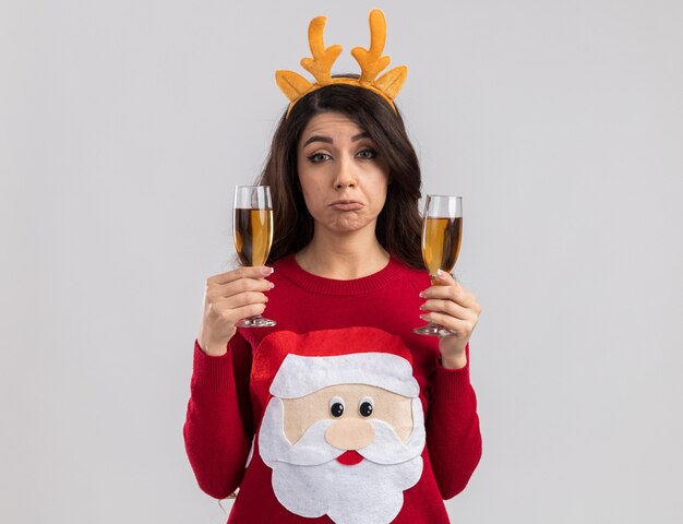 Clueless young pretty girl wearing reindeer antlers headband and santa claus sweater holding two glasses of champagne looking 