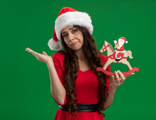 Clueless young pretty girl wearing santa hat holding santa on rocking horse figurine looking at camera showing empty hand isolated on green background