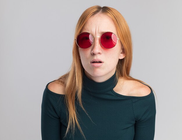 Clueless young redhead ginger girl with freckles in sun glasses looking at camera on white