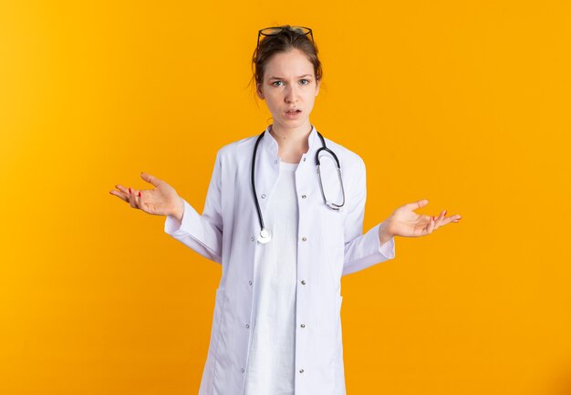 Clueless young slavic girl in doctor uniform with stethoscope keeping hands open isolated on orange wall with copy space