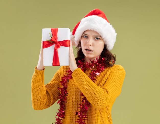 clueless young slavic girl with santa hat and with garland around neck holding christmas gift box isolated on olive green background with copy space