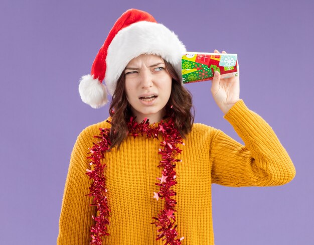 Clueless young slavic girl with santa hat and with garland around neck holds paper cup close to ear isolated on purple background with copy space