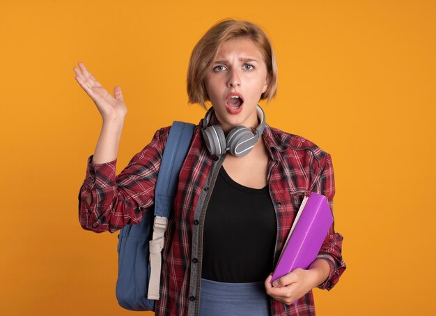 Clueless young slavic student girl with headphones wearing backpack stands with raised hand holds book and notebook 