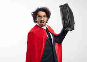 Foto gratuita il giovane supereroe senza tracce in vetri ottici che indossa la tuta con mantello rosso tiene la borsetta in pelle isolata sul muro bianco