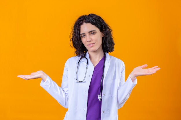 Clueless young woman doctor wearing white coat with stethoscope shrugging shoulders looking uncertain and confused having no answer spreading palms standing on isolated orange