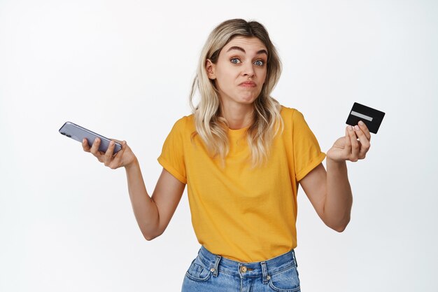 Clueless young woman holds smartphone and credit card, shrugging shoulders with puzzled face expression on white.