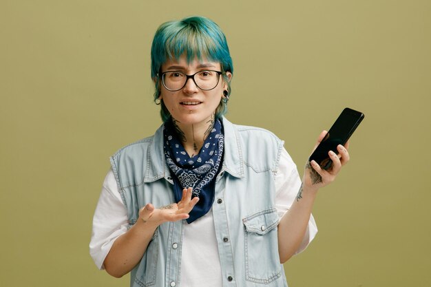 올리브 녹색 배경에 격리된 카메라를 쳐다보는 빈 손을 보여주는 휴대전화를 들고 목에 안경 반다나를 착용한 단서 없는 젊은 여성