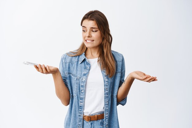 Clueless young woman with blond hair and tattoos, shrugging, staring at smartphone puzzled, standing against white wall